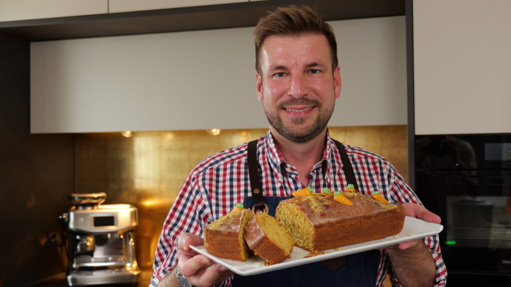 Georg hält einen länglichen Teller mit einem angeschnittenen Käsekuchen in der Hand. Im Hintergrund sieht man die Küche.